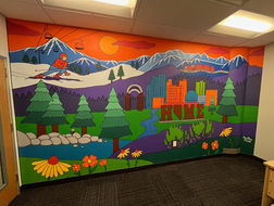 Mural with skier, snowy mountains, pine trees, the Spark Victorian  Square sign, the Reno Home sign, Reno city skyline, Truckee river, Lake Tahoe, and flowers.