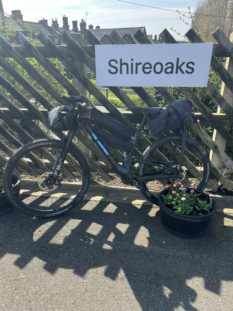 Bicycle with travel bags against a wooden fence at Shireoaks station. A planter with colorful flowers is nearby. Bright, clear day.