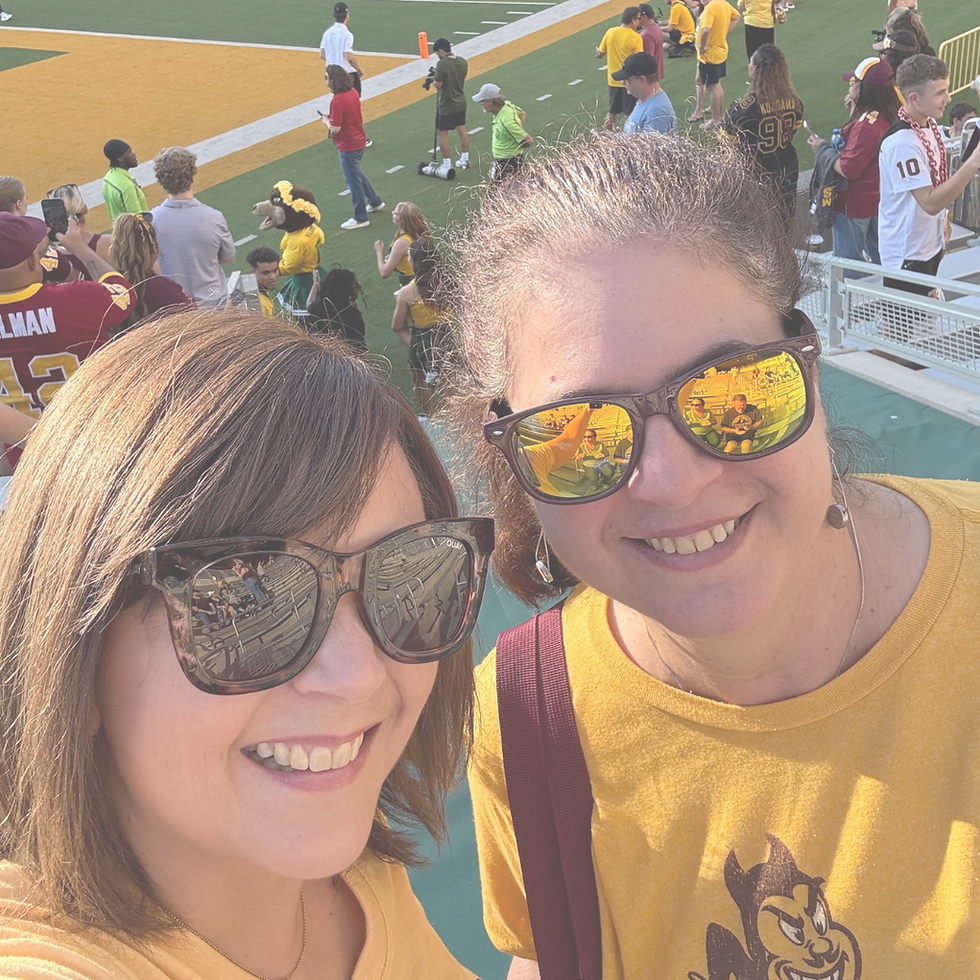 Two women in sunglasses and gold shirts smile for a selfie at a sports event. Fans are visible in the stadium background.