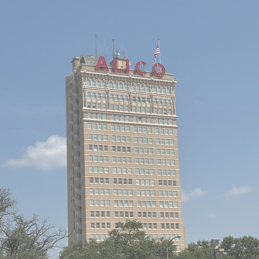 Tan-bricked skyscraper with "ALICO" atop, American flag, and blue sky background. Urban setting with green trees in front.