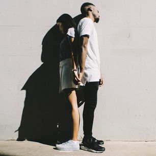 Two people stand back-to-back, holding hands against a sunlit wall. The shadows are prominent, creating a contemplative mood.