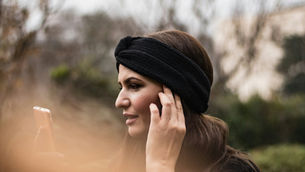 Woman in a black leather jacket uses a smartphone outside on a sunny day. Blurred city street and benches in the background.