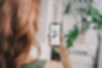 Woman holding a smartphone showing a review of "Oatly" milk. Indoor hallway with plants. Rainbow tattoo on her arm. Calm setting.