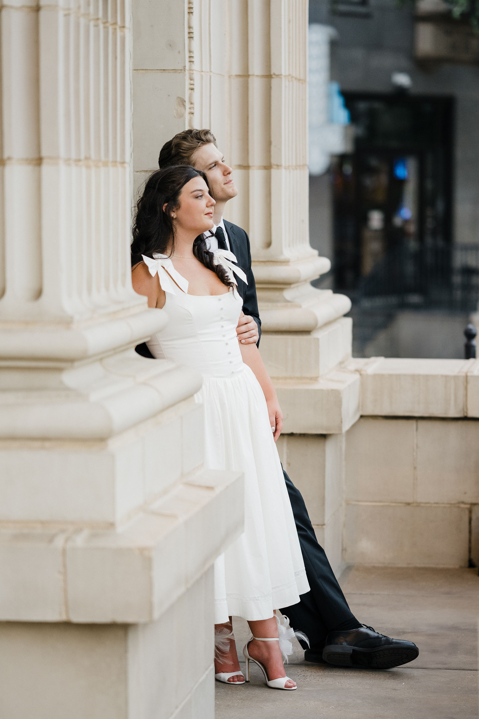 Engagement photo of a couple leaning against stone columns with urban backdrop, modern editorial Dallas engagement photography with romantic city vibes.