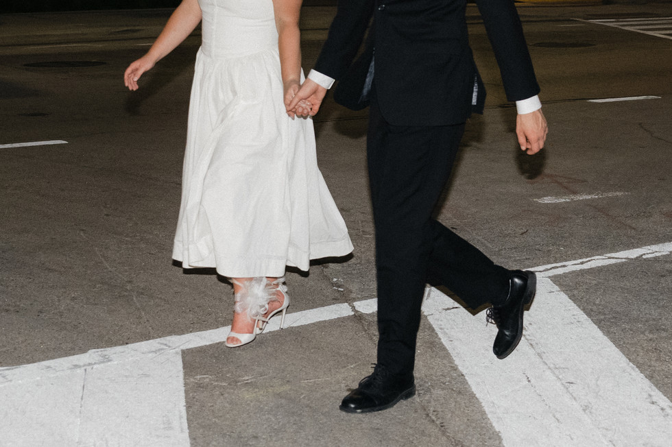 Bride in a white dress and groom in a black suit holding hands and walking across a crosswalk during a modern Dallas engagement session.