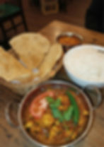 Indian meal on a wooden table: spicy curry in a metal dish, white rice in a bowl, papadum in a basket, and orange chutney in a small bowl.