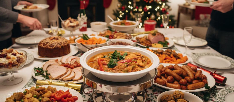 Festive dinner scene with various dishes on a table, Christmas tree lit in the background, and people serving themselves, creating a warm holiday mood.