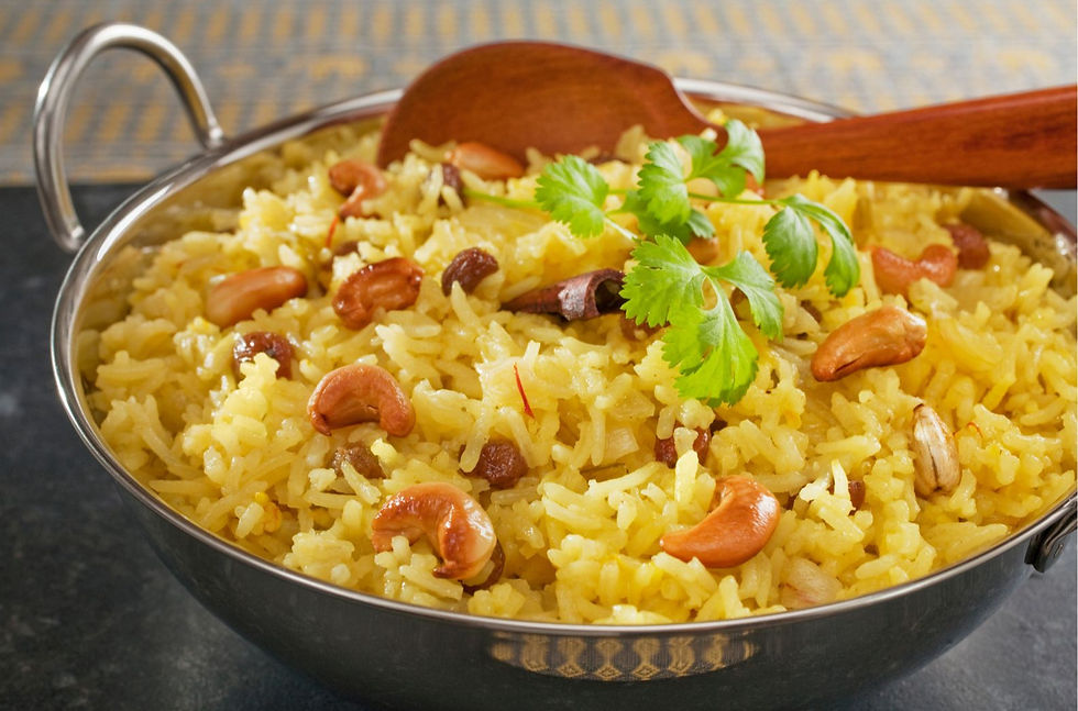 Yellow pilau rice with cashews and raisins in a metal bowl, garnished with cilantro. A wooden spoon rests on top, set on a dark surface.