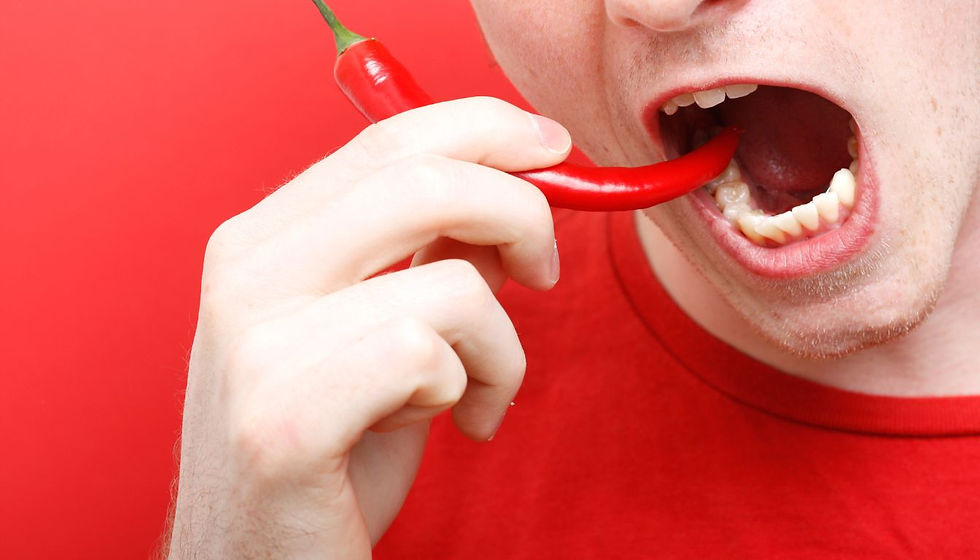 Close-up of a person in a red shirt biting a bright red chili pepper against a red background, with an intense expression.