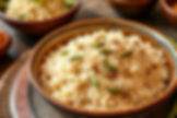 Bowl of seasoned rice with green herbs on top, placed on a wooden table. The background shows blurred bowls, creating a warm ambiance.