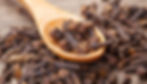 A wooden spoon filled with dried cloves rests on a wooden surface, surrounded by more cloves. The image is in warm, earthy tones.
