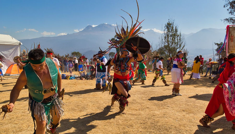 Colorful Indigenous dancers in feathered costumes perform outdoors on a dusty field with mountains and tents behind.