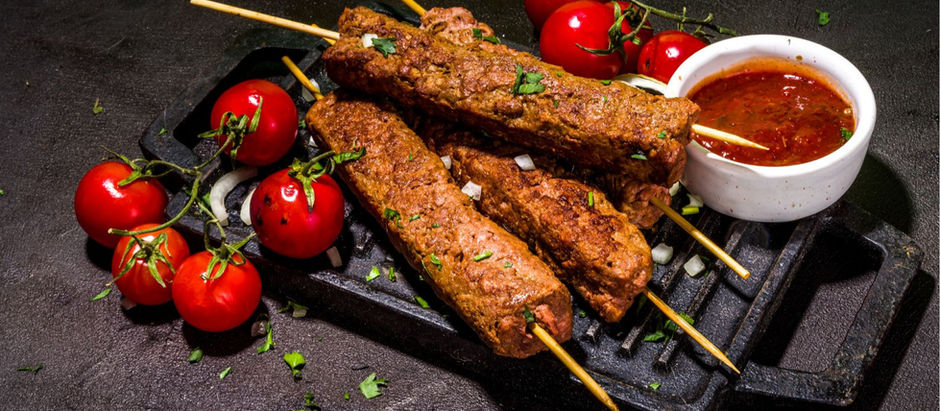 Grilled hassina kebabs on skewers atop a black grill platter with vine tomatoes, a white bowl of red sauce, and chopped herbs on a dark surface.