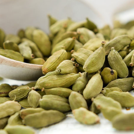 Green cardamom pods spill from a wooden scoop onto a white surface, creating a natural and earthy setting with soft lighting.
