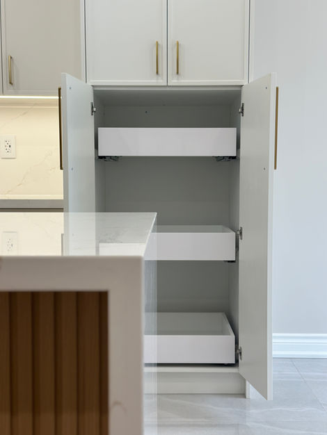 Pantry with open doors, drawers, and shelving unit above, blurred island in the foreground, natural wood tones, white kitchen in the background.