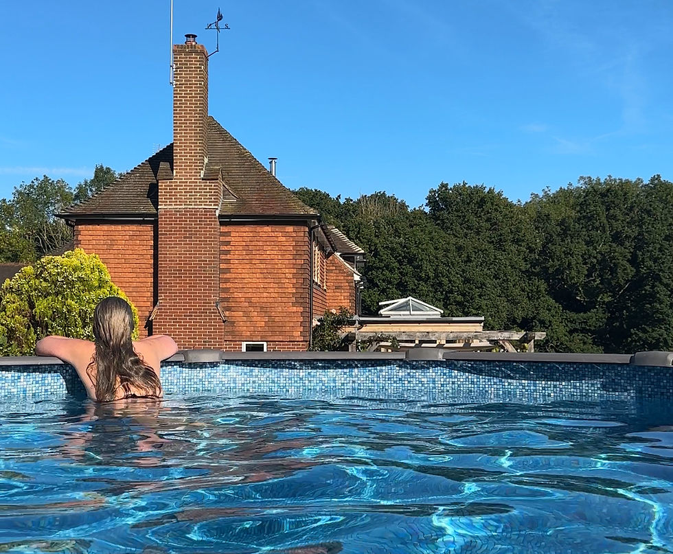 Woman with long hair relaxes at pool edge, facing a red brick house. Bright blue water sparkles under clear sky. Trees surround the scene.