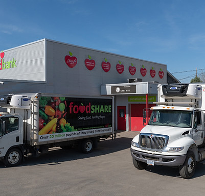 The grey exterior of the kamloops food bank building is shown on a sunny day with two 5 tonne trucks parked in front. One of the trucks has a colourful food share logo and images of fresh fruits and vegetables on the side of the truck.