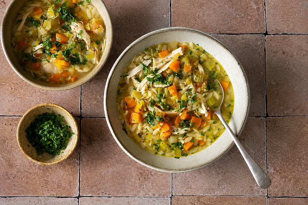 Overhead horizontal photo of two bowls of homemade chicken noodle soup with shredded chicken, orzo pasta, carrots, celery, herbs, and broth on a rustic tiled surface, with a small bowl of fresh chopped parsley beside them.