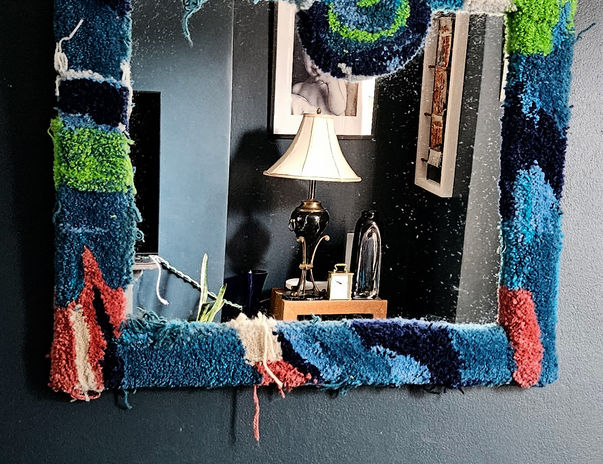 Sidh Tufted Mirror by Ryka Design displayed on dark navy wall showing square format with hand-tufted frame in teal, navy and green Axminster wool with distinctive circular swirl motif