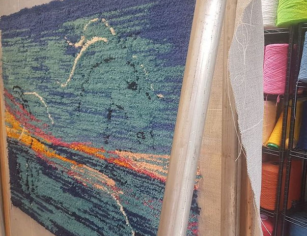 Sea Canter Tufted Rug by Ryka Design shown on tufting frame displaying complete seascape composition in deep navy, teal and cobalt blue with vivid amber, orange and red horizon band in hand-tufted wool pile with colourful yarn spools on studio shelving