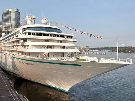 Medium size cruise ship white with aqua blue trim in the port of Vancouver with mountains, trees and sky rises in the background