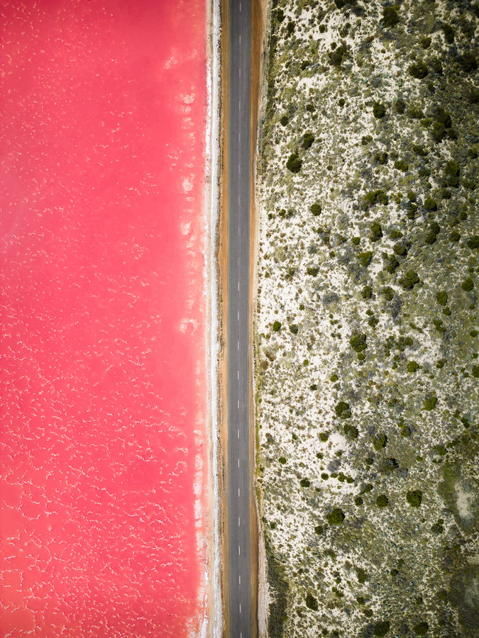Drohnenaufnahme der Hutt Lagoon (Pink Lake) in Australien aus einem 90-Grad-Winkel nach unten, aufgenommen von Kyburz Visuals. In der Mitte verläuft eine gerade Strasse, links der leuchtend pinke See und rechts Sand und grünes Gebüsch.