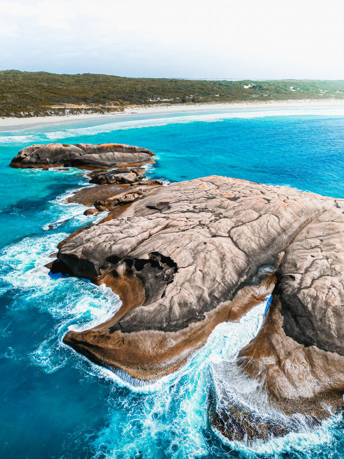 Luftaufnahme von Twilight Beach, Australien. Im Vordergrund sind große, von Wellen umspülte Felsen im türkisblauen Wasser zu sehen. Im Hintergrund erstreckt sich ein langer, weißer Sandstrand mit einer grünen, hügeligen Landschaft.