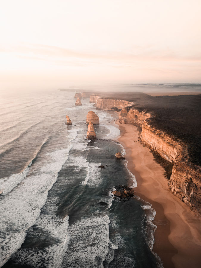 Luftaufnahme der 12 Apostles an der Great Ocean Road in Australien bei Sonnenuntergang. Die Felsformationen erheben sich aus dem Wasser entlang der Küste, während die Wellen an den Strand rollen. Das warme, sanfte Licht der untergehenden Sonne taucht die Szene in pastellfarbene Töne und verstärkt die natürliche Schönheit der Kalksteinfelsen und der rauen Küstenlinie.