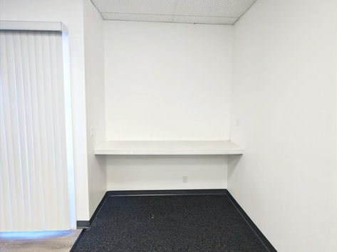An empty office space with a gray carpet, a white floating shelf on the wall, and a large white sliding door on one side.