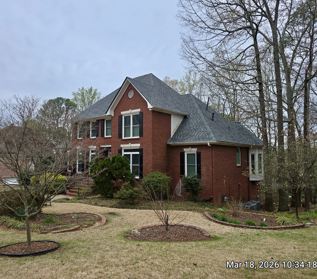 Side view of a large two-story red brick residential house with a grey roof, surrounded by green trees and a front yard
