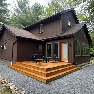 Exterior view of the completed Pocono Pines home renovation by Robert Ace Builders, showcasing the new custom sunroom addition, expansive energy-efficient windows, oversized wood deck, updated entry door, and thoughtful design that complements the home's existing T1-11 siding and wooded mountain setting.