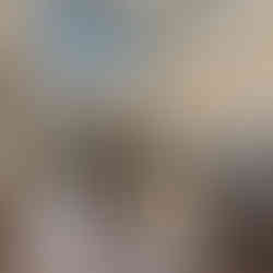 Open-concept living area featuring dark wood flooring, light neutral walls, and a vaulted ceiling with exposed wood beams. A stone fireplace is positioned between glass doors and large black-trimmed windows that fill the space with natural light and views of the surrounding trees. The living area flows into a kitchen with white cabinetry, stainless steel appliances, pendant lighting, and a contrasting dark island, while a seating arrangement defines the lounge space.