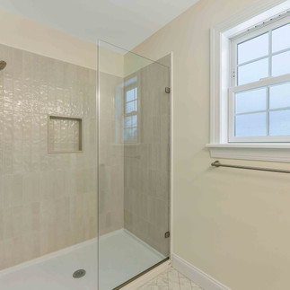 Interior view of a glass-enclosed walk-in shower with light tile walls, a built-in shower niche, chrome fixtures, marble-look tile flooring, and a nearby window bringing natural light into the bathroom.