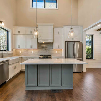 Kitchen featuring white cabinetry, stainless steel appliances, and a large contrasting island with a light countertop beneath two pendant lights. A farmhouse-style sink is positioned below a window, complemented by a tile backsplash and under-cabinet lighting. Dark wood flooring continues throughout the space, and an adjacent dining area is visible through the open doorway.