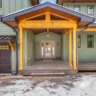 Close-up exterior view of the front entry of a custom home in Lake Naomi, PA featuring green siding, natural wood posts and beams, and a covered porch with a peaked roof and wood-paneled ceiling. A glass-paneled front door is centered beneath the entryway and flanked by wall-mounted lantern-style lights. The porch connects to an attached garage with a wood door and black metal awning, and snow-covered ground surrounds the front of the home.