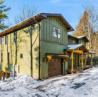 Angled exterior front view of a custom two-story home in Lake Naomi, PA featuring green vertical and horizontal siding, a dark metal roof with extended eaves, and natural wood trim. The home includes an attached single-car garage with a wood door and black metal awning, a covered front porch supported by exposed wood posts, and multiple rectangular windows along both levels. The house sits on a sloped, wooded lot with a snow-covered driveway and mature trees surrounding the property.