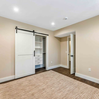 Unfurnished interior room with beige walls, white trim, and dark wood flooring partially covered by a textured area rug. A sliding barn door opens to a built-in closet with shelving and drawers, while an open doorway provides access to an adjacent space.