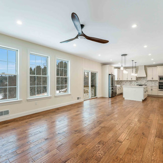 Interior view of an open-concept living area with hardwood flooring, light cream walls, recessed lighting, a modern ceiling fan, and multiple large windows that fill the space with natural light. The adjoining kitchen with white cabinetry and center island is visible in the background.