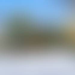 Exterior front view of a fully remodeled two-story home in Lake Naomi, PA featuring green vertical and horizontal siding, a dark metal roof, and natural wood trim. The home includes an attached single-car garage with a wood door and black metal awning, a covered front porch supported by exposed wood posts, and a recessed entryway with a glass-paneled front door. Large rectangular windows line both levels of the home. The house sits on a wooded lot with snow-covered ground and mature trees surrounding the property.