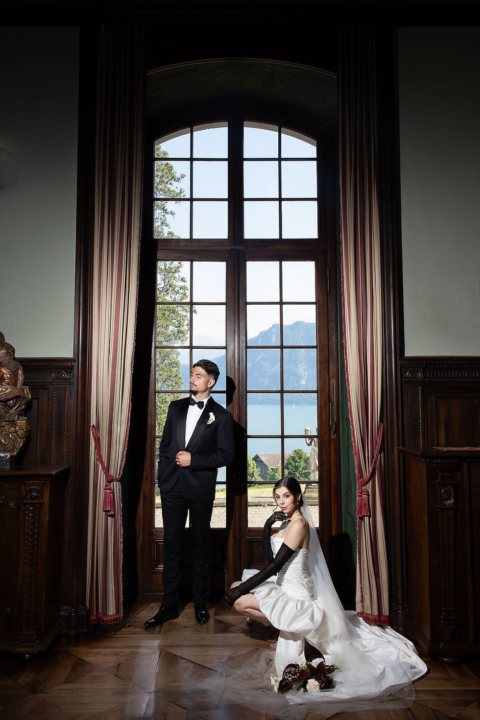 Bride in white gown and groom in black tux pose by a tall window with lake and mountains beyond, in a stately room