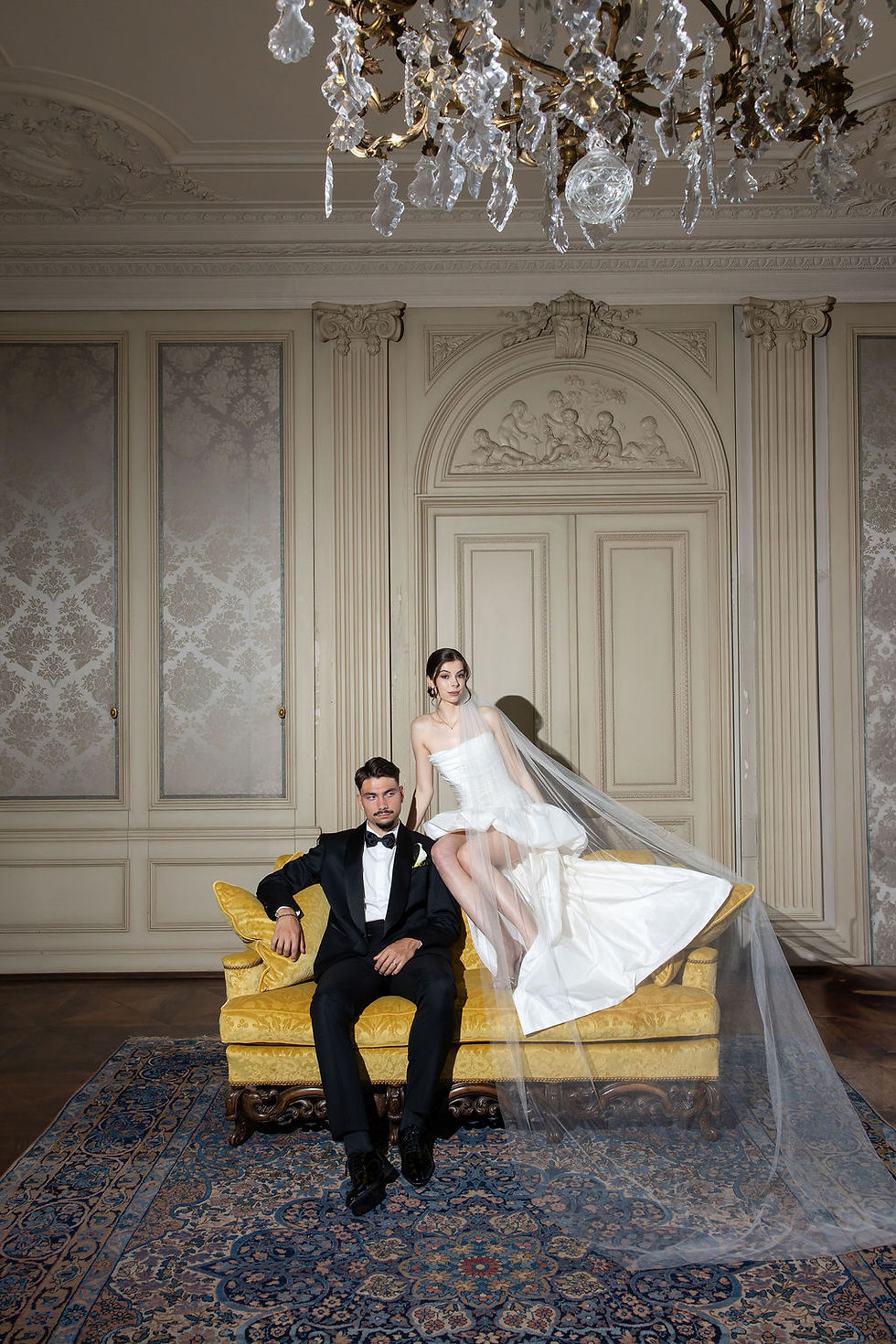 Bride in white gown and veil sits on a yellow sofa beside groom in a black tuxedo in an ornate room with chandelier.