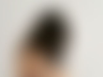 A woman with long dark hair, seen in profile and partially nude, hugs herself gently while holding a small bouquet of dried lavender against her chest. Her face is obscured by her hair, and the background is a plain, light-colored wall.