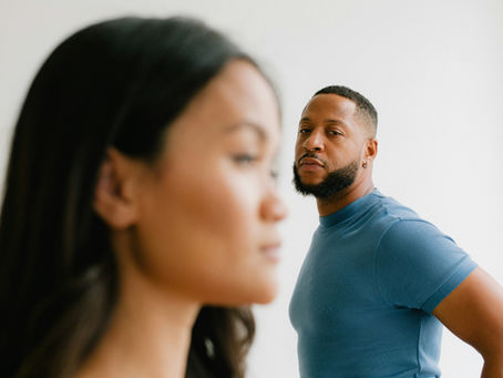 A Black man in a blue shirt stands in focus, looking directly at the camera with a serious expression. In the foreground, an East Asian woman is out of focus, facing sideways and partially obscuring the man. The background is minimal and white, emphasizing the subjects' expressions and body language.