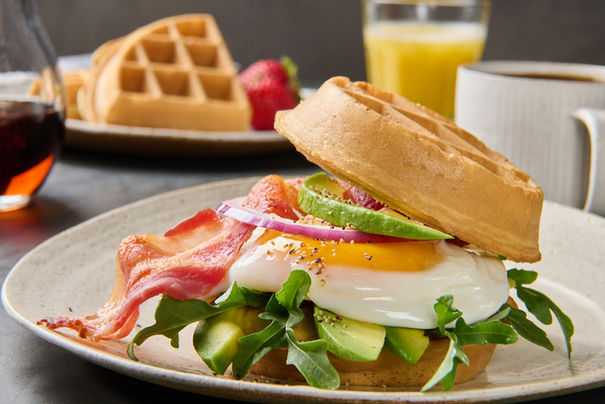 Close-up of a waffle breakfast sandwich with bacon, egg, avocado, arugula, and red onion on a plate with waffles and orange juice in the background.