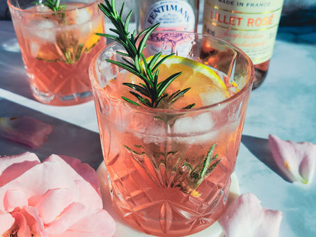 Rosa Getränk mit Rosenlimonade, Lillet Rose, Zitronenscheibe und Rosmarin in Glas, daneben Rosenblüten. Im Hintergrund Flaschen mit Aufschrift „Rose Lemonade“.