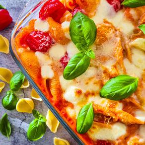 Nudelauflauf mit Tomaten, Mozzarella und Basilikum in einer Auflaufform, daneben Nudeln und Kirschtomaten auf Steinfläche