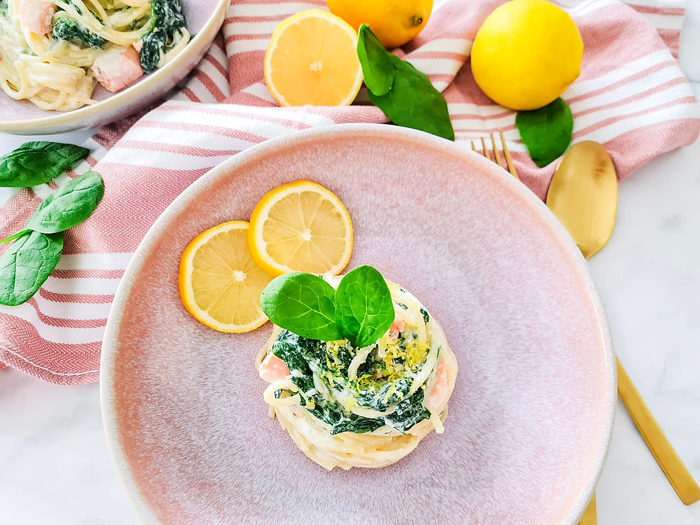 Pasta mit Spinat und Lachs auf rosa Teller, mit Zitronenscheiben und grünem Blatt, daneben goldene Gabel und Löffel.