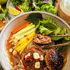 Große Schüssel mit vietnamesischer Bun Cha: Fleischbällchen, Gemüse und Kräutern; Stäbchen, Limetten und Schälchen auf Holztisch.