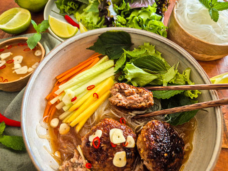Große Schüssel mit vietnamesischer Bun Cha: Fleischbällchen, Gemüse und Kräutern; Stäbchen, Limetten und Schälchen auf Holztisch.