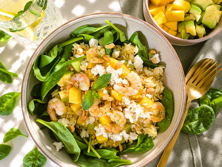 Bunte Bowl mit Spinat, Reis Nudeln, Garnelen, Mango, Avocado, Feta und Nüssen auf hellem Tisch, sonnig und frisch.